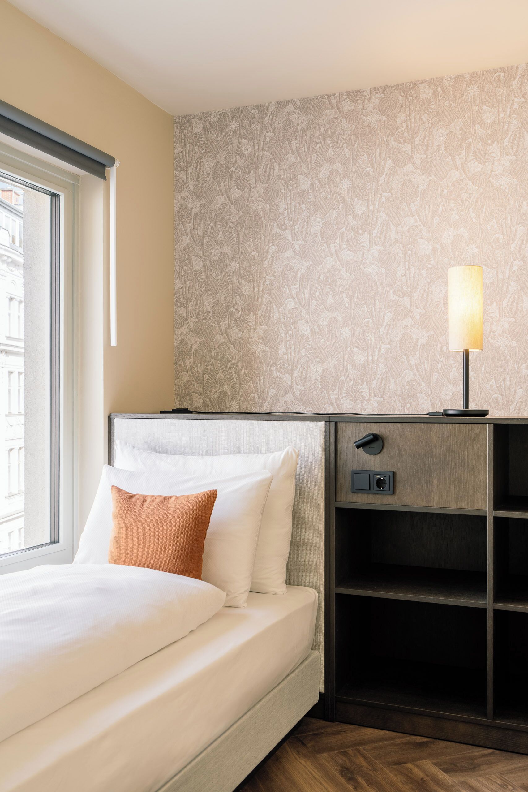 A hotel room with a bed, a nightstand, a lamp, and a window.