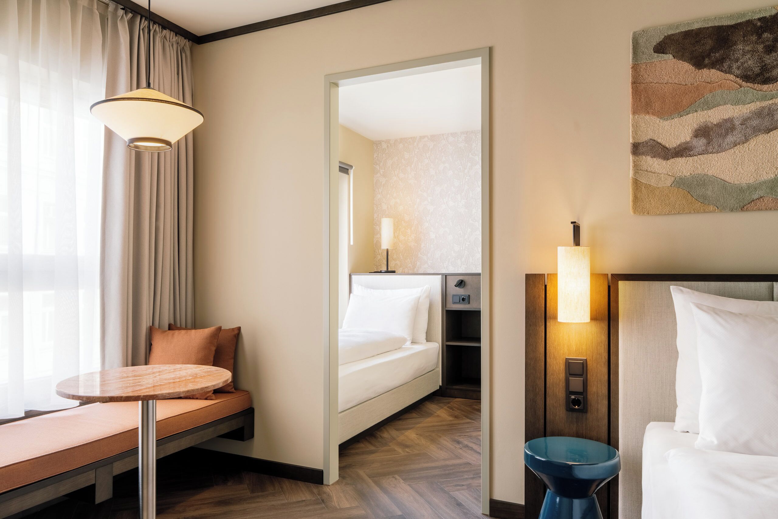 A hotel room with a bed, a seating area with a table, a mirror, a wall-mounted lamp, and a painting.