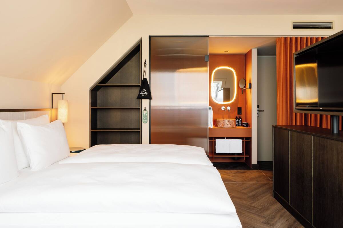 A hotel room with a bed, a TV, a bathroom with a mirror, and a shelf with toiletries.