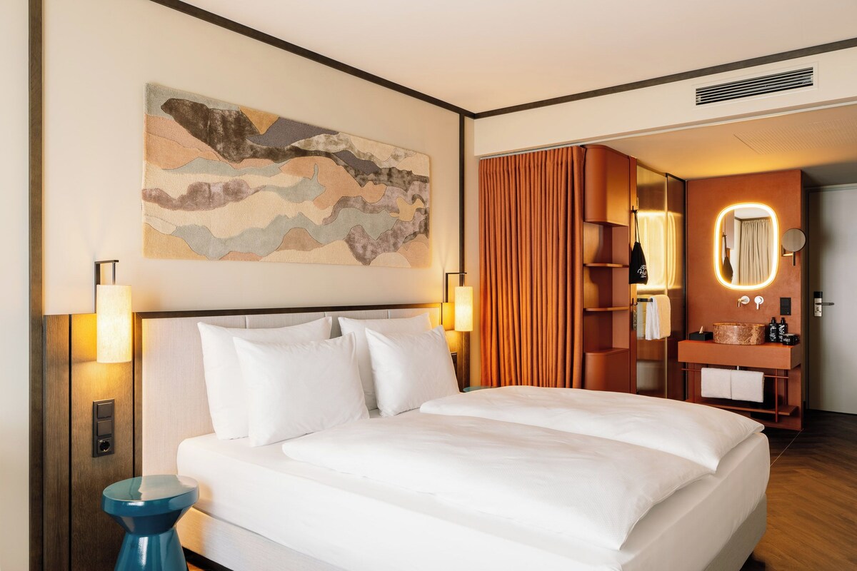 A hotel room with a large bed, a wall-mounted artwork, a wooden wardrobe, a bathroom with a mirror, and a blue side table.