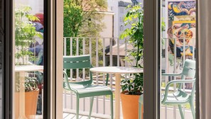 A room with a large window, a balcony with chairs and potted plants, and a view of buildings and trees.