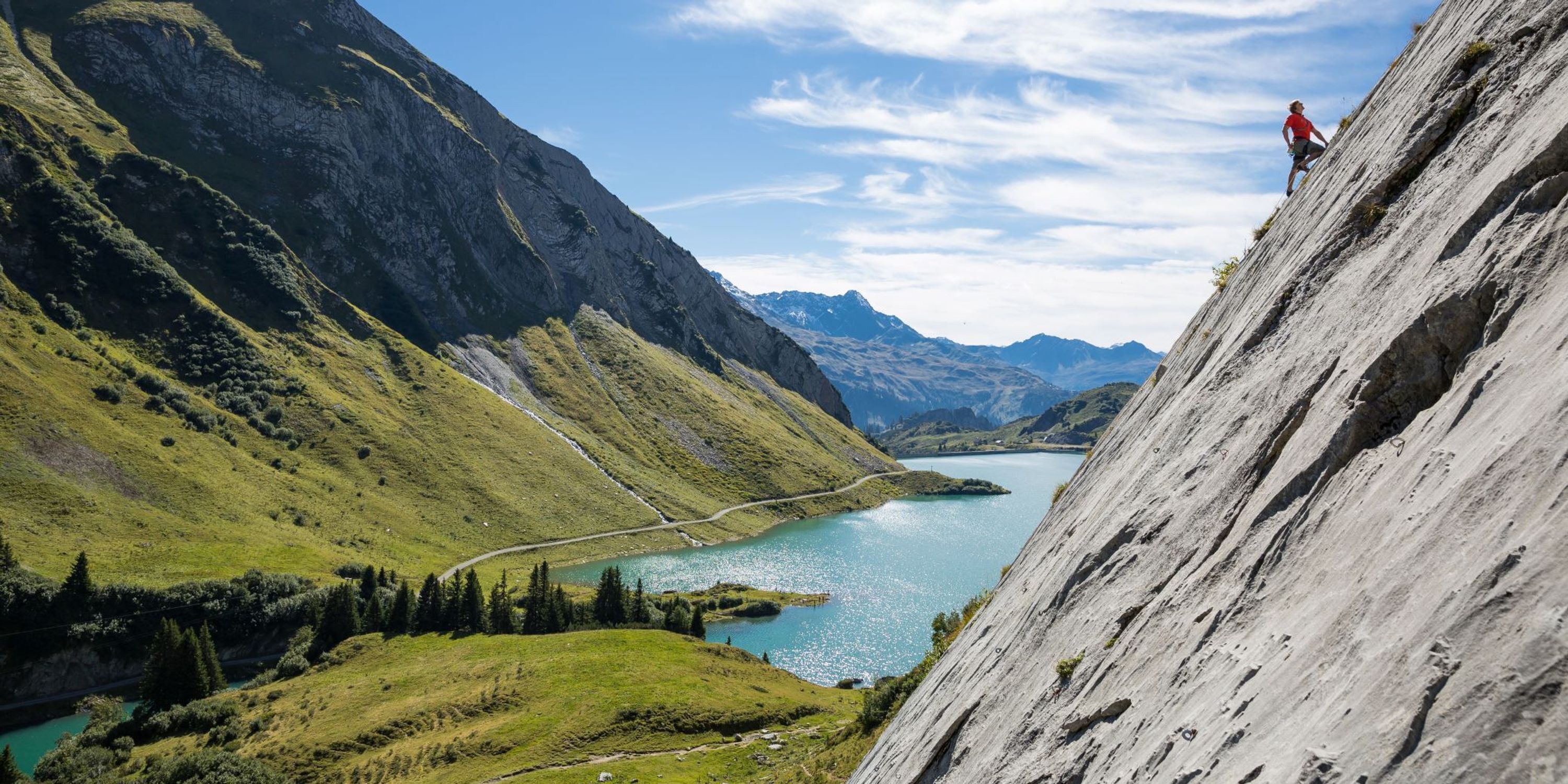 Un paysage montagneux avec un lac, une route et une personne qui escalade une paroi rocheuse escarpée.