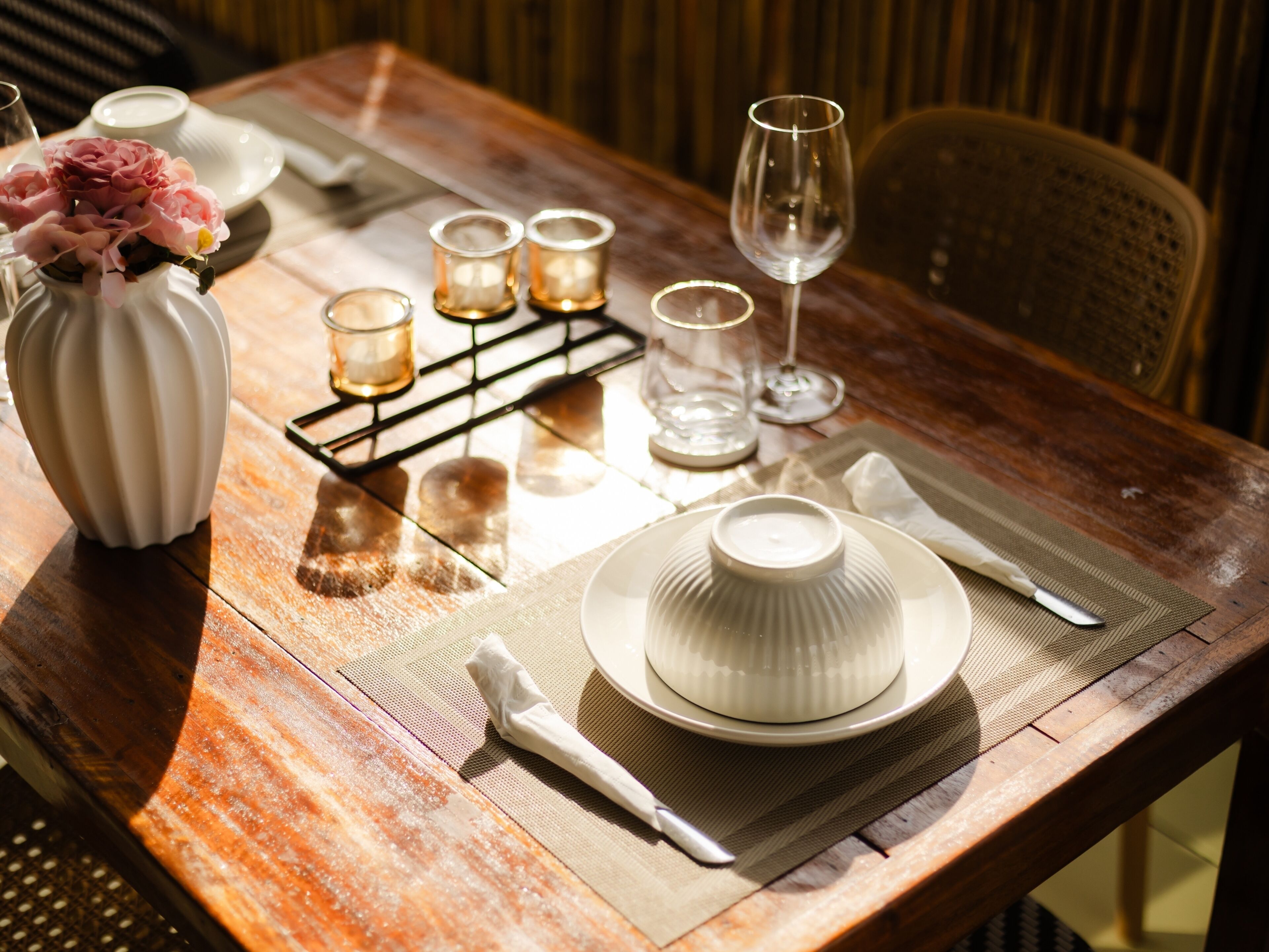 Una mesa de madera dispuesta con platos blancos, un jarrón de flores rosas y velas.