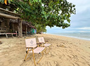 A beach with two folding chairs under a tree, a wooden structure, and a beach hut in the background. - L1 Lanta Resort (Ko Lanta)