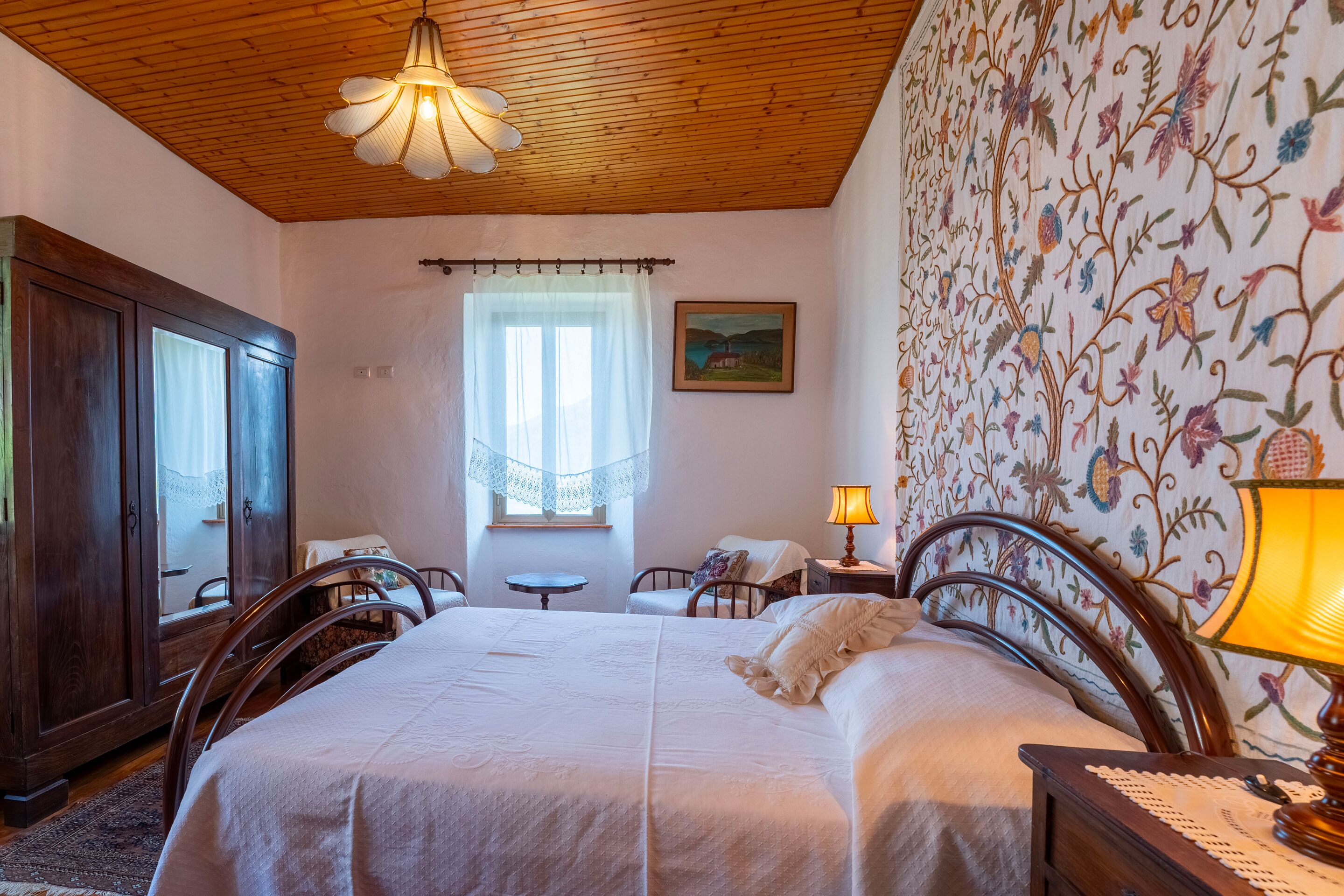 Un dormitorio con techo de madera, una cama con ropa de cama blanca, una mesita de noche con lámpara, un armario de madera, una ventana con cortina de encaje y un papel tapiz floral.