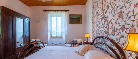 Un dormitorio con techo de madera, una cama con ropa de cama blanca, una mesita de noche con lámpara, un armario de madera, una ventana con cortina de encaje y un papel tapiz floral.