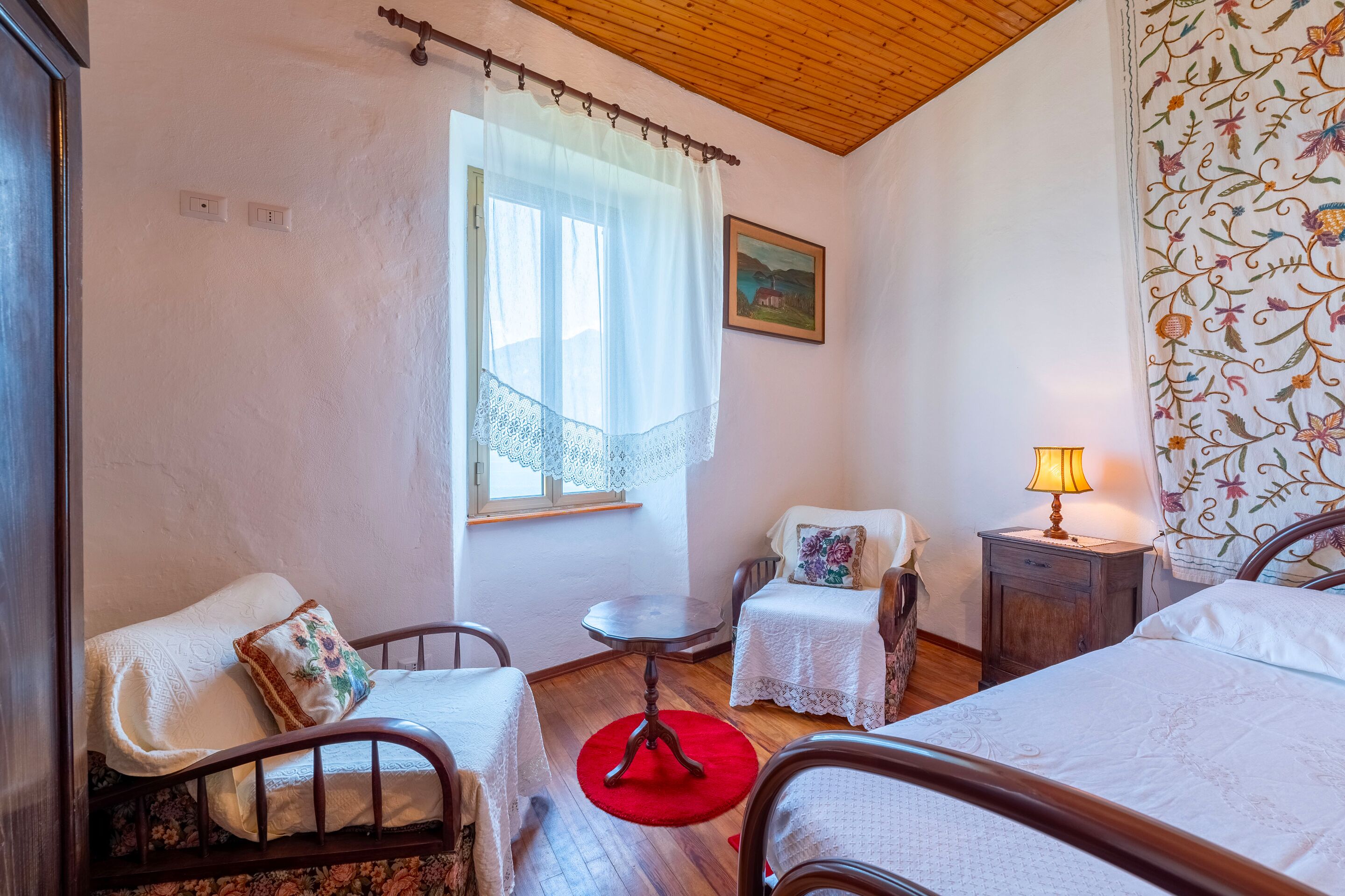 A room with a bed, two chairs, a small table, a lamp, a window with a curtain, and a painting on the wall.