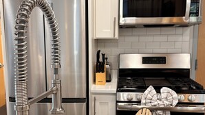 Una cocina con armarios blancos, electrodomésticos de acero inoxidable y un recipiente de cerámica con manchas que contiene utensilios de madera.