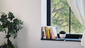 Una cama bien hecha con ropa blanca, una planta en maceta, un estante con libros y una ventana con vistas a árboles.