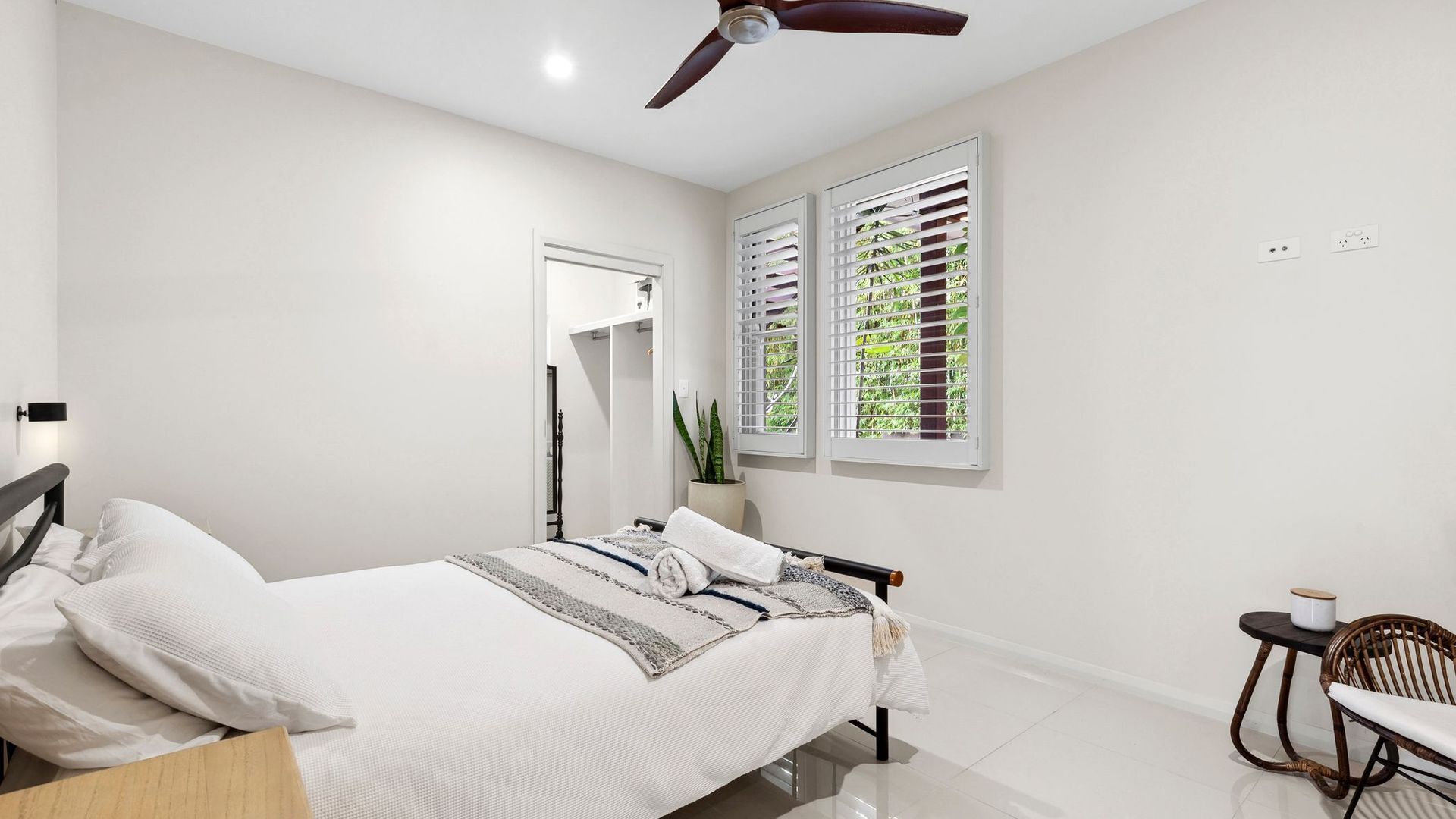 A modern bedroom with a bed, a ceiling fan, two windows with shutters, a small table, and a chair.