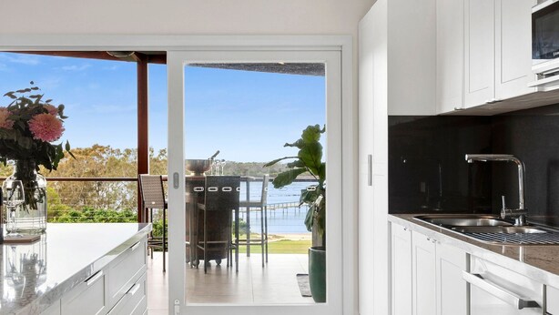 A modern kitchen with a view of the sea, a dining area with outdoor seating, and a large window. - Urunga Magic – A beautiful waterfront retreat in Urunga with serene river and ocean views (Urunga)