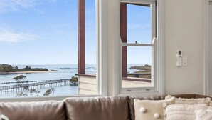A room with a large window overlooking a coastal area, featuring a wooden pier and a sandy beach. - Urunga Magic – A beautiful waterfront retreat in Urunga with serene river and ocean views (Urunga)
