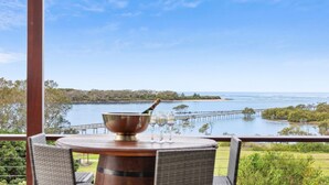 A table with a copper bowl, glasses, and a bottle on a balcony overlooking a coastal landscape with a pier. - Urunga Magic – A beautiful waterfront retreat in Urunga with serene river and ocean views (Urunga)