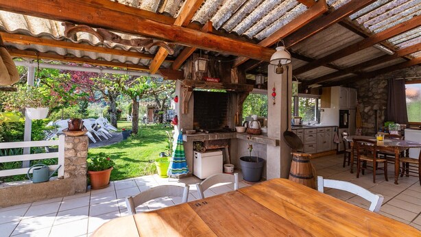 An outdoor patio with a wooden table and chairs, a stone wall, and a grill area.