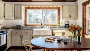 A kitchen with white cabinets, a large window, a round wooden table with a fruit platter, wine glasses, and a vase with flowers. - The Sugarcreek Farmhouse•Hot Tub|Theater|Fire Pit (Blue Ridge)