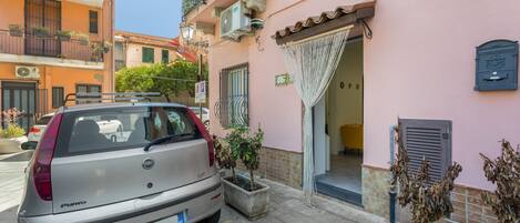 A parked silver car in a narrow street with a pink building, a mailbox, and a door with a curtain.
