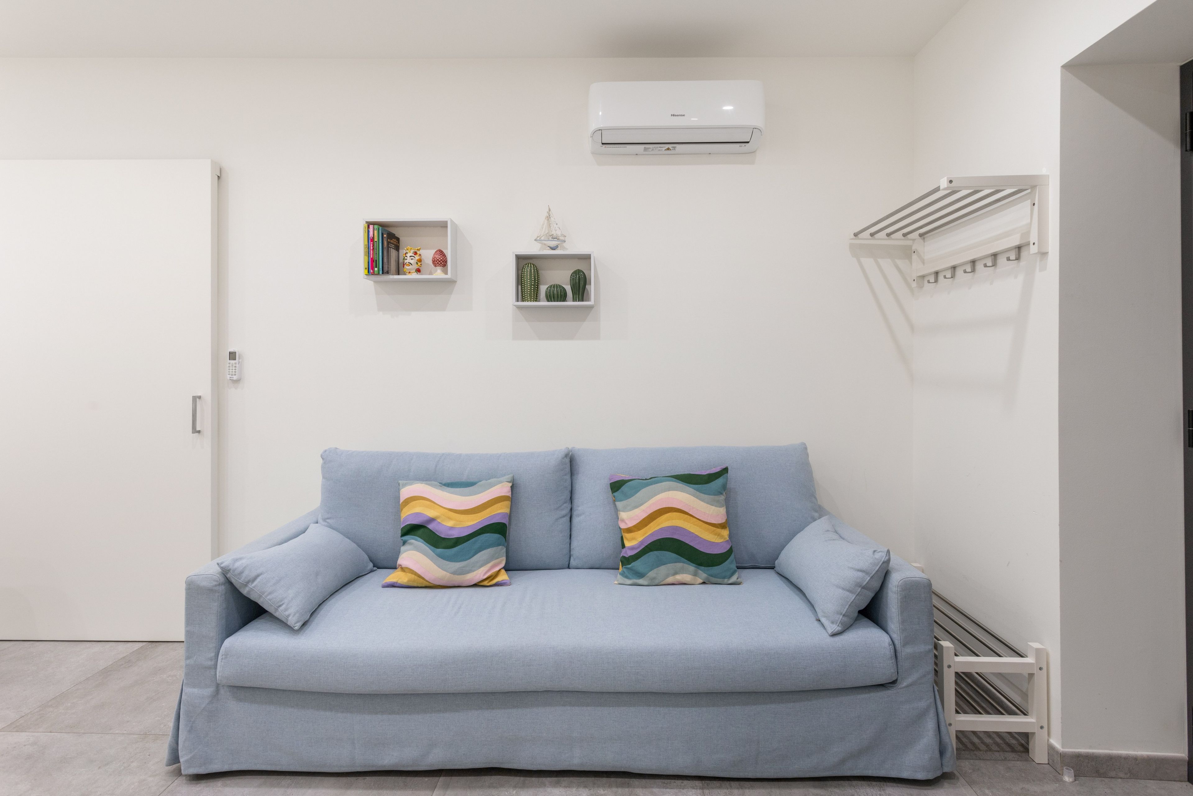A light blue sofa with two colorful pillows, a white air conditioning unit, and a small shelf with decorative items.