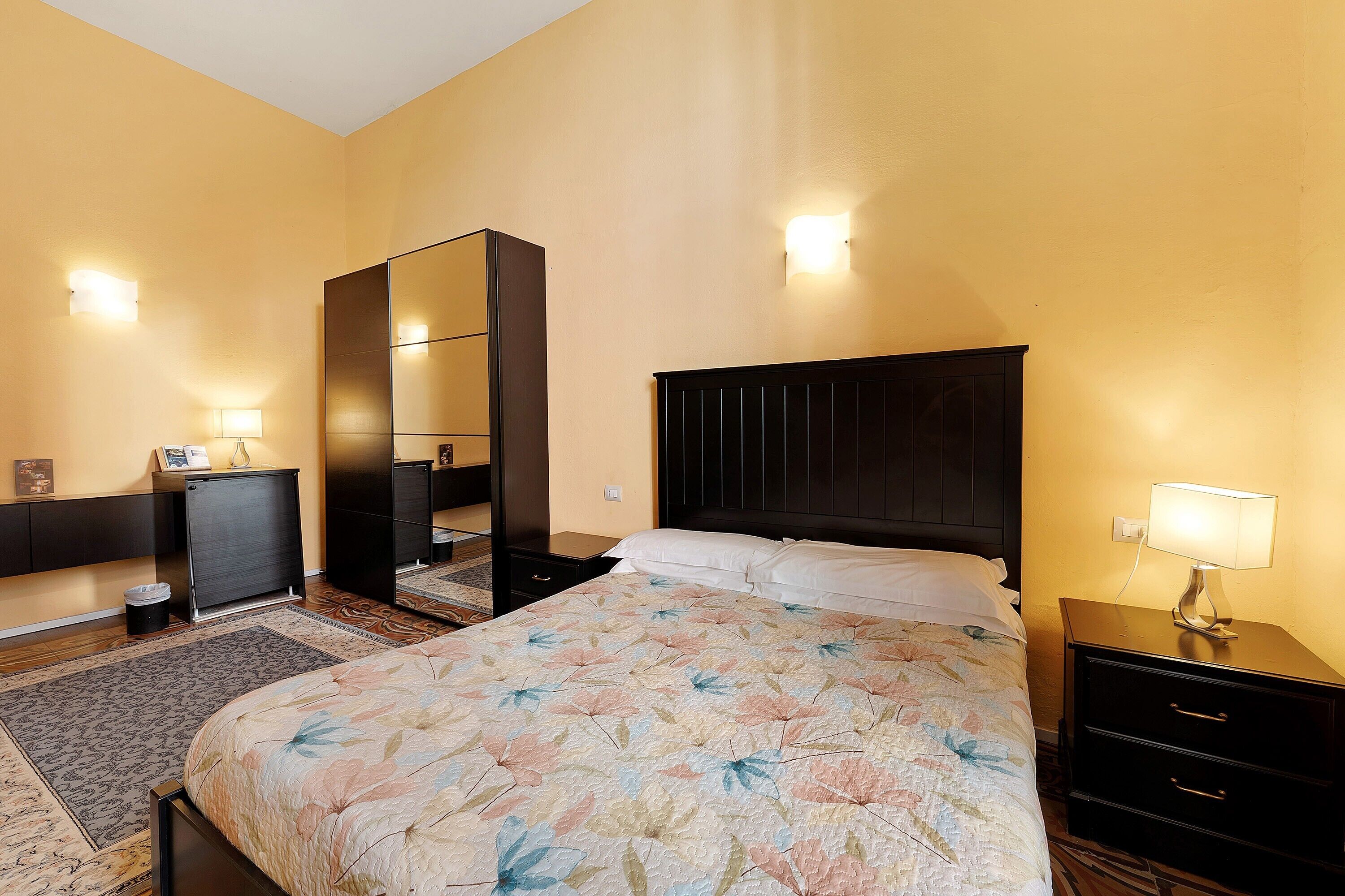 A hotel room with a bed, a nightstand, a dresser, a mirror, and a wall-mounted light.