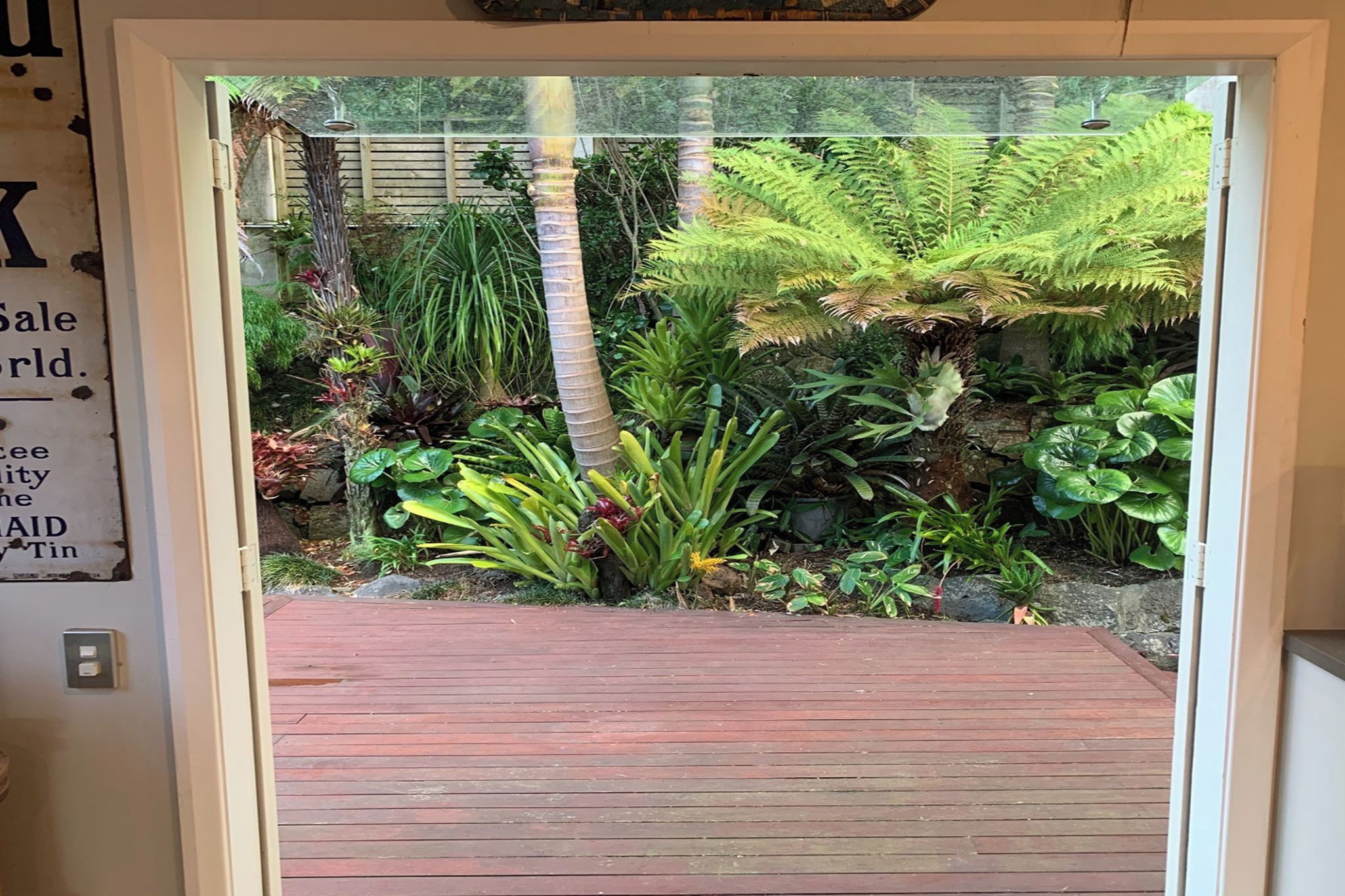 Une terrasse en bois mène à un jardin luxuriant où l’on trouve diverses plantes, et un panneau est fixé au mur.
