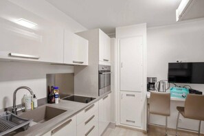 A compact kitchen with white cabinetry, a built-in oven, a coffee maker, and a small seating area with a chair.