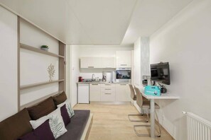 A compact living space with a kitchenette, a sofa, a TV, and a small table with a welcome box.