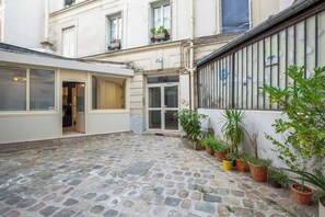 A courtyard with a stone floor, a door leading to a room, and several potted plants.