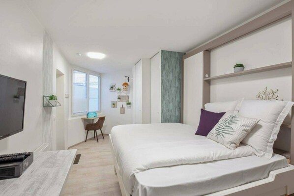 A bedroom with a bed, a desk, a chair, a TV, and a window.
