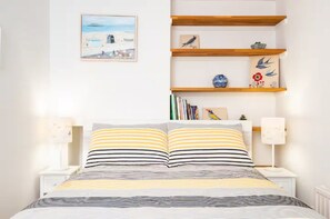 Un dormitorio con una cama, dos mesitas de noche, un aparador, un estante con libros y objetos decorativos, y un cuadro en la pared.