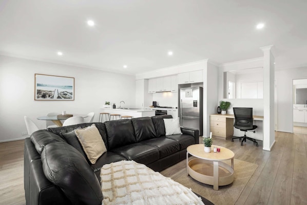 A modern living room with a black leather sofa, a round coffee table, and a dining area with a white table and chairs. - Cherry St Retreat, Armstrong Creek (Armstrong Creek)