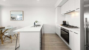A modern kitchen with white cabinetry, a central island, stainless steel appliances, and a dining area with a glass table and chairs. - Cherry St Retreat, Armstrong Creek (Armstrong Creek)