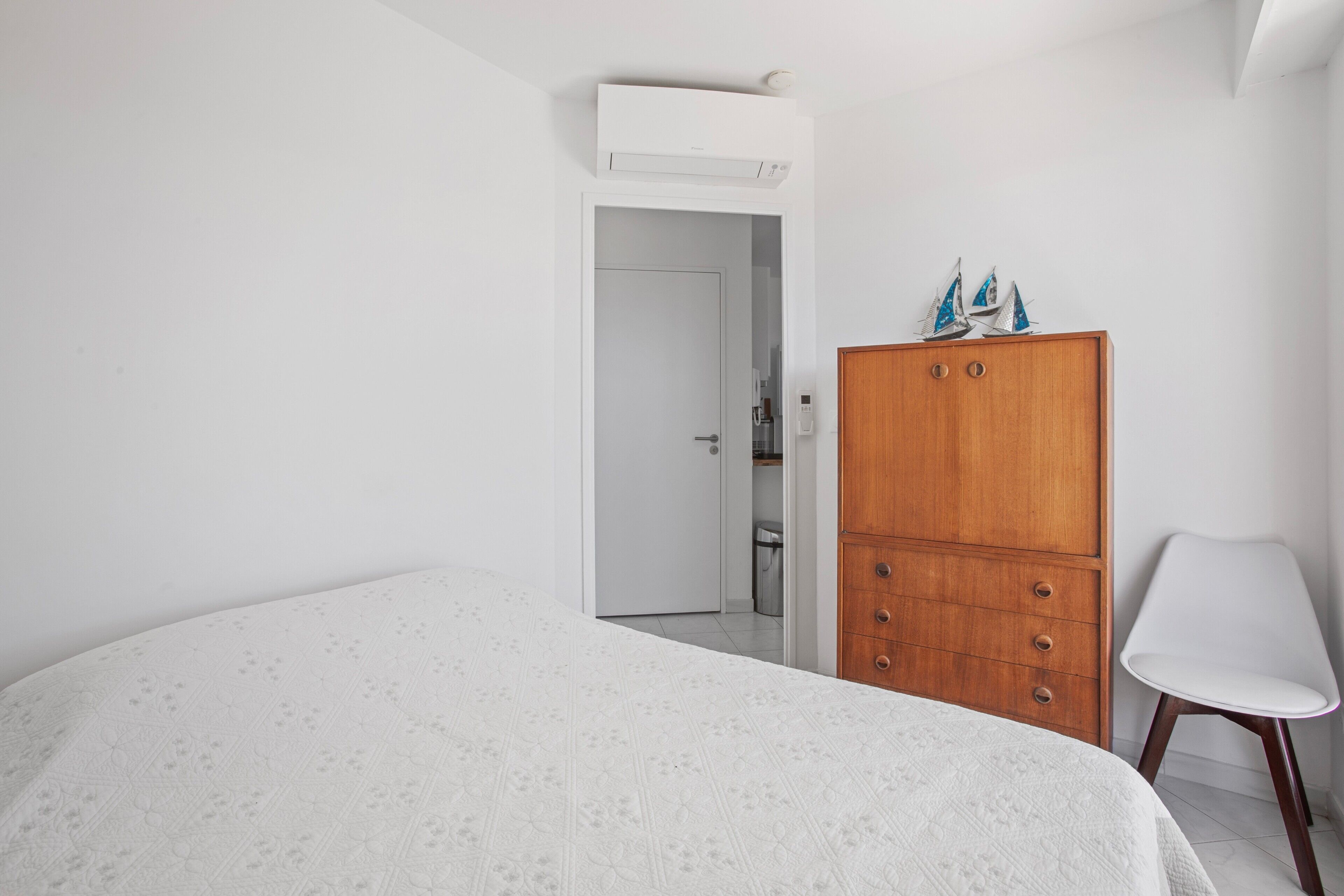 Une chambre à coucher comprenant un lit, une armoire en bois, une chaise blanche et une porte donnant sur une salle de bain.
