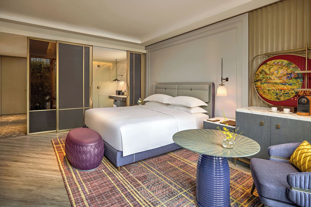 A modern hotel room with a large bed, a sitting area with a table, and a view of the outdoors.