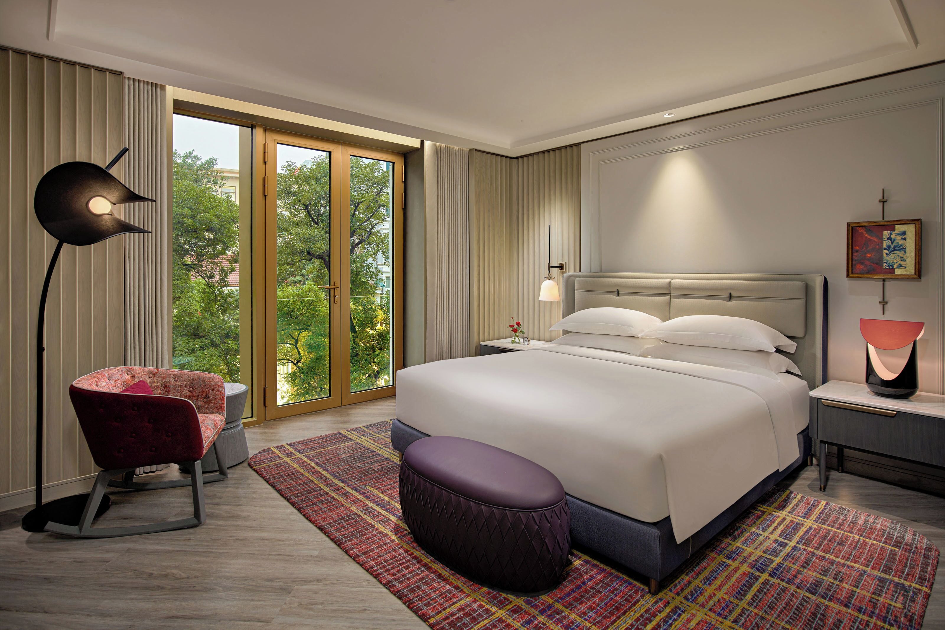 Ein modernes Hotelzimmer mit einem großen Bett, einem roten Sessel, einer Stehlampe und Blick auf Grün.