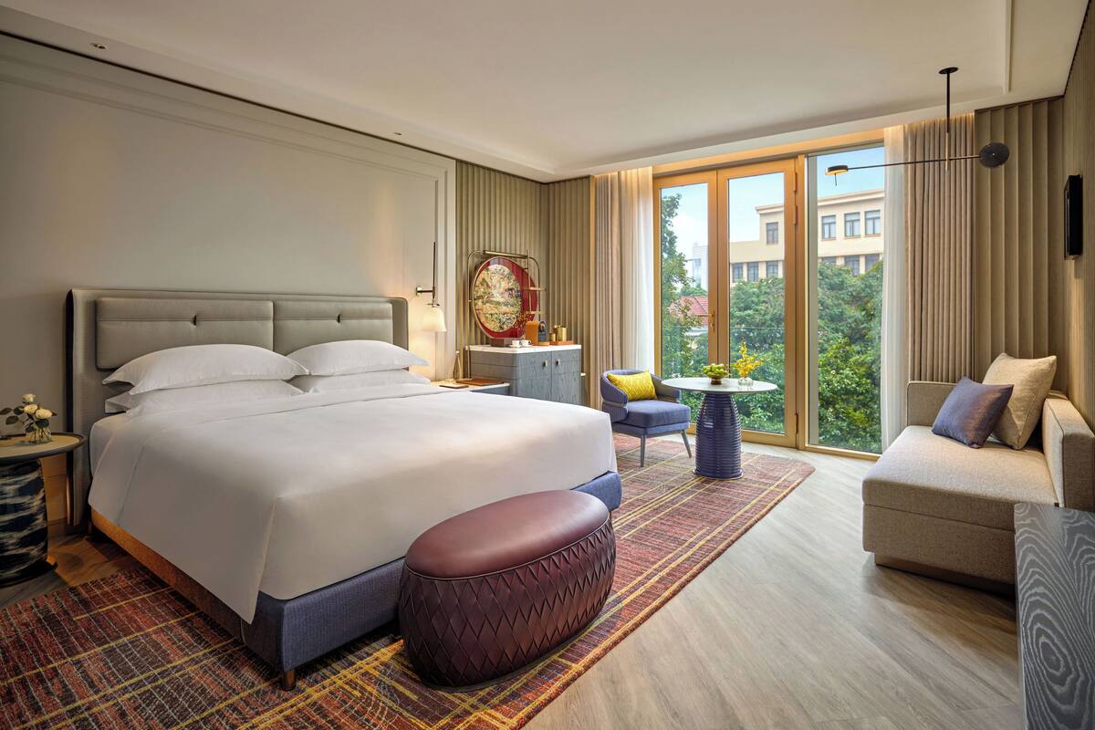 A hotel room with a large bed, a sofa, a small table, and a view of greenery outside.