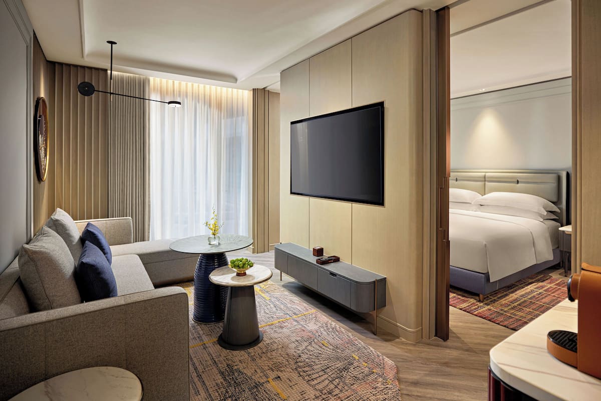 A modern hotel room with a sofa, a small round table with a vase, a flat-screen TV mounted on the wall, a bed with white linens, and a patterned rug.