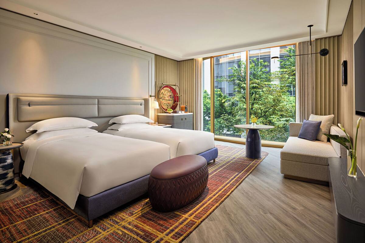 A modern hotel room with a large bed, a sofa, a small table, and a view of greenery outside.