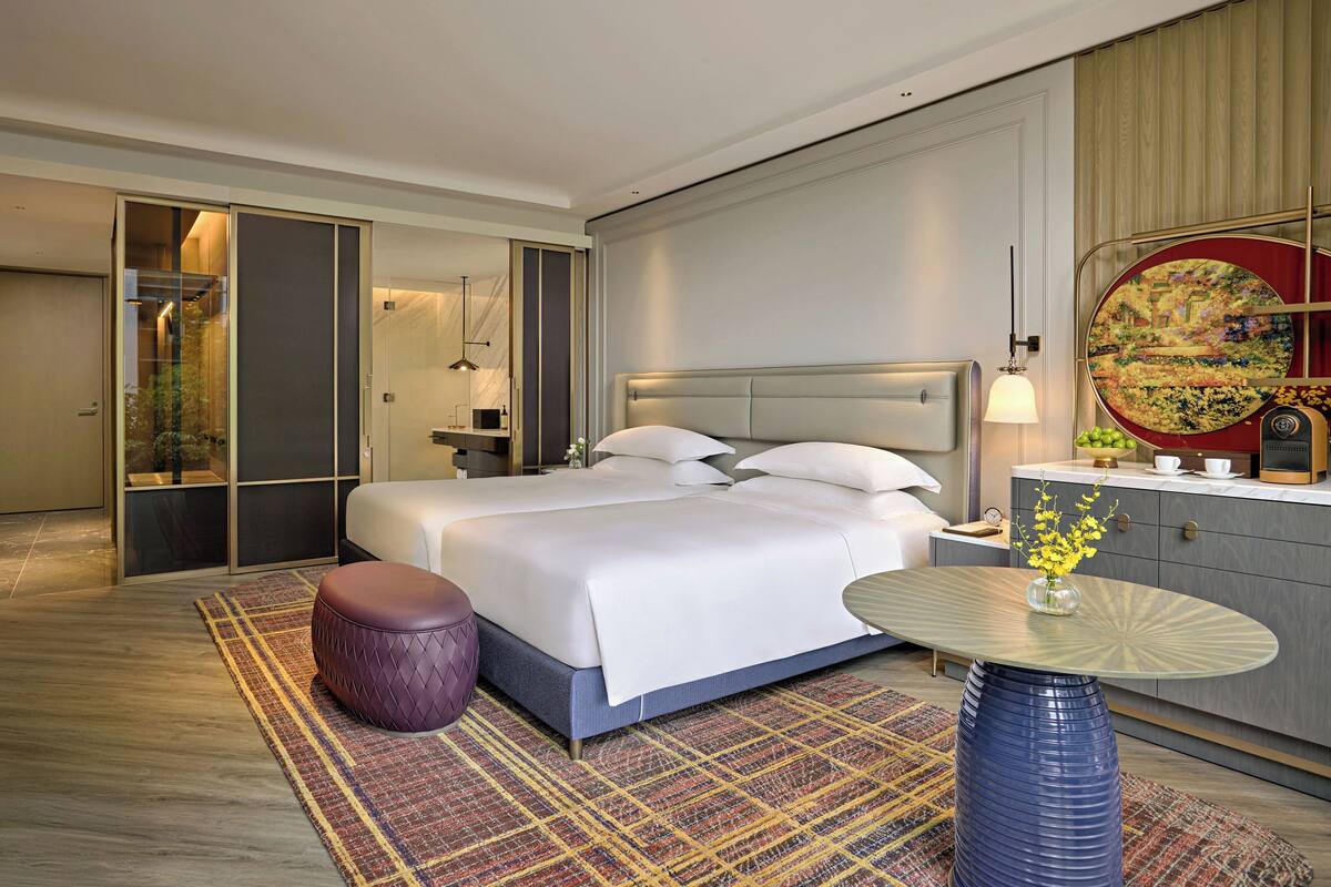 A modern hotel room with a large bed, a round table with a vase of flowers, a purple ottoman, and a large mirror on the wall.