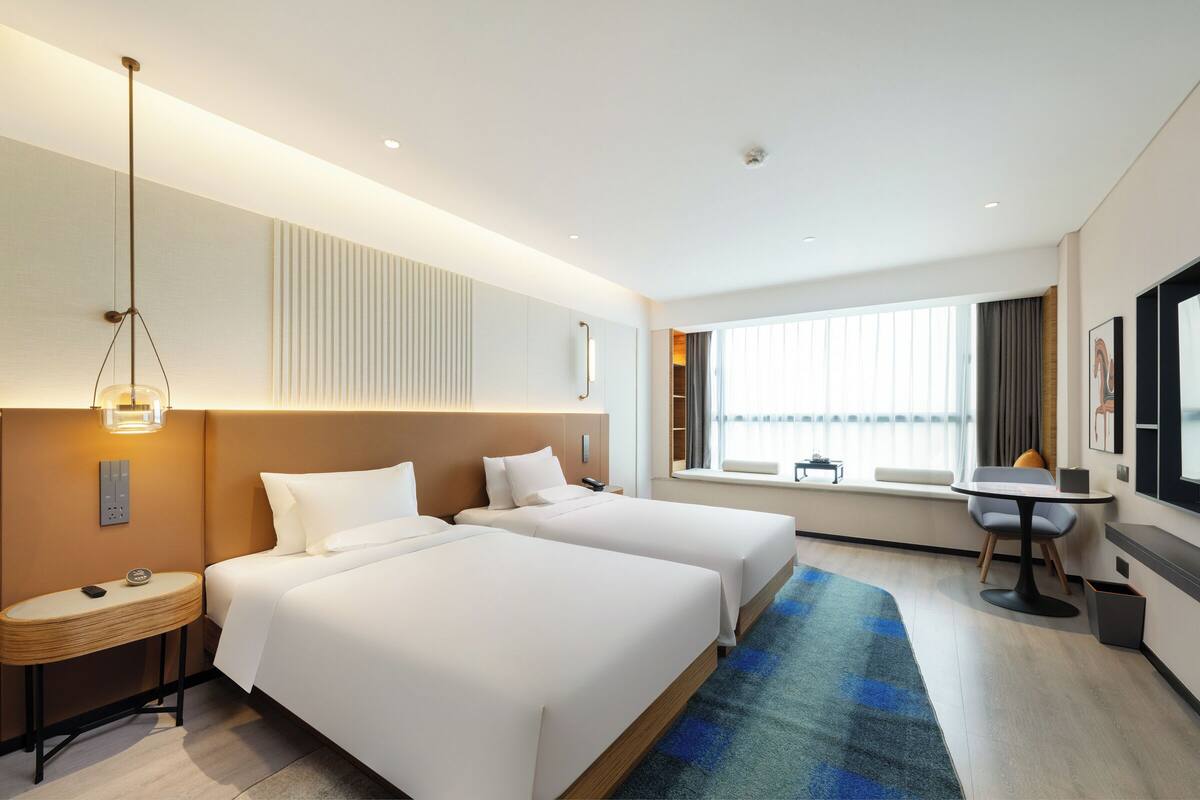 Een hotelkamer met twee bedden, een klein tafeltje, een stoel, een bureau en een groot raam met gordijnen.