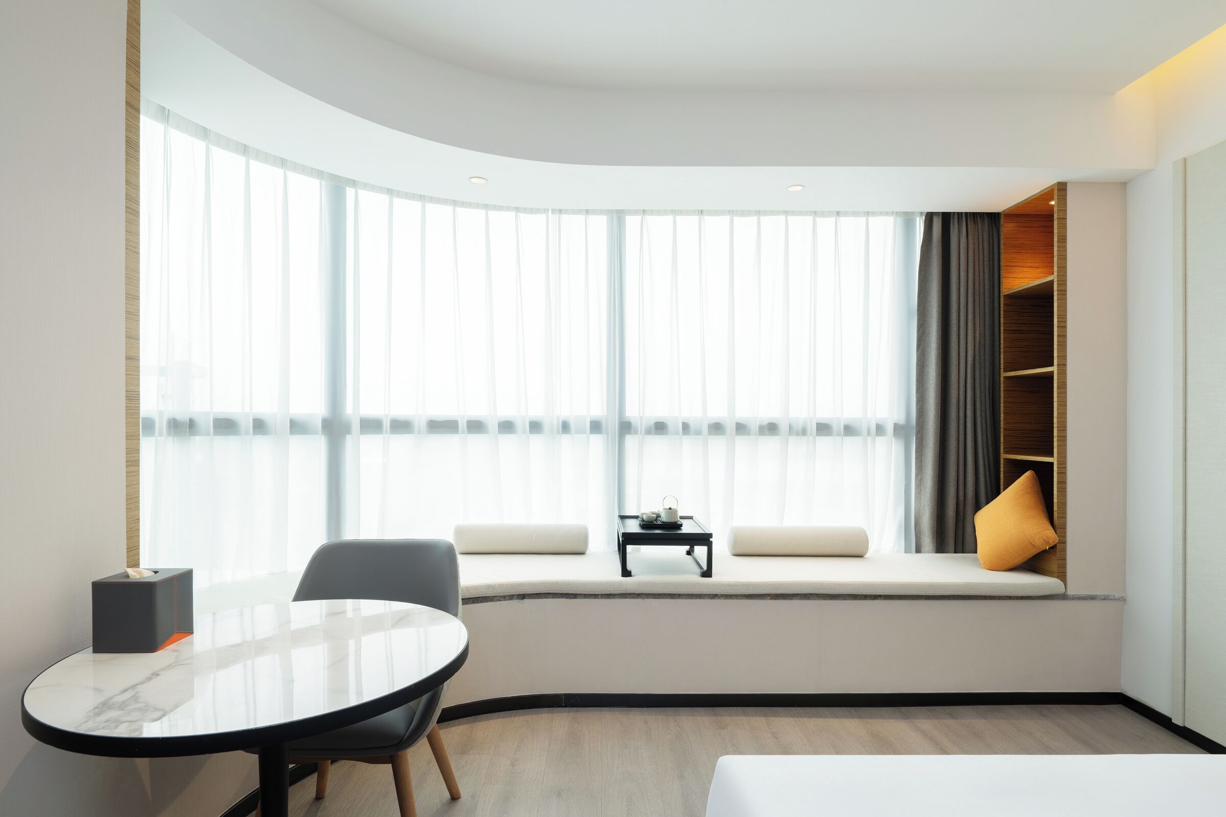 A modern hotel room with a curved ceiling, a round table with chairs, a sofa, a small table with a lamp, and a large window with curtains.