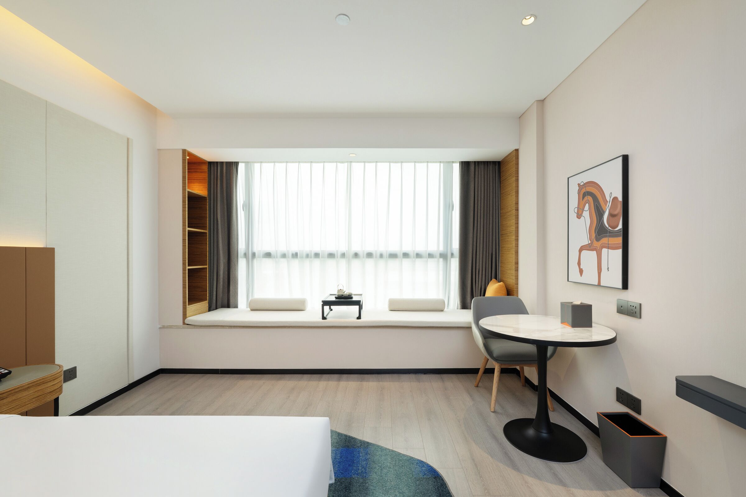 A modern hotel room with a large window, a bed, a small table, a chair, and a desk.