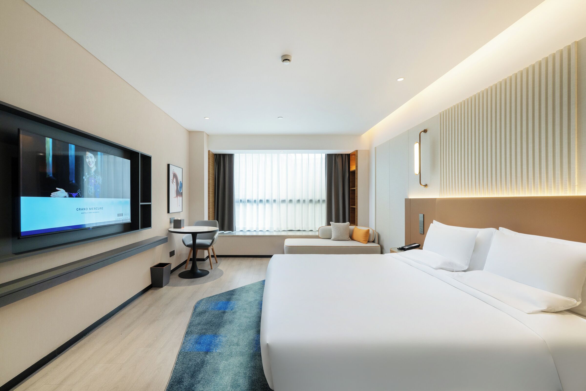 A modern hotel room with a large bed, a flat-screen TV, a small table, and a window with curtains.
