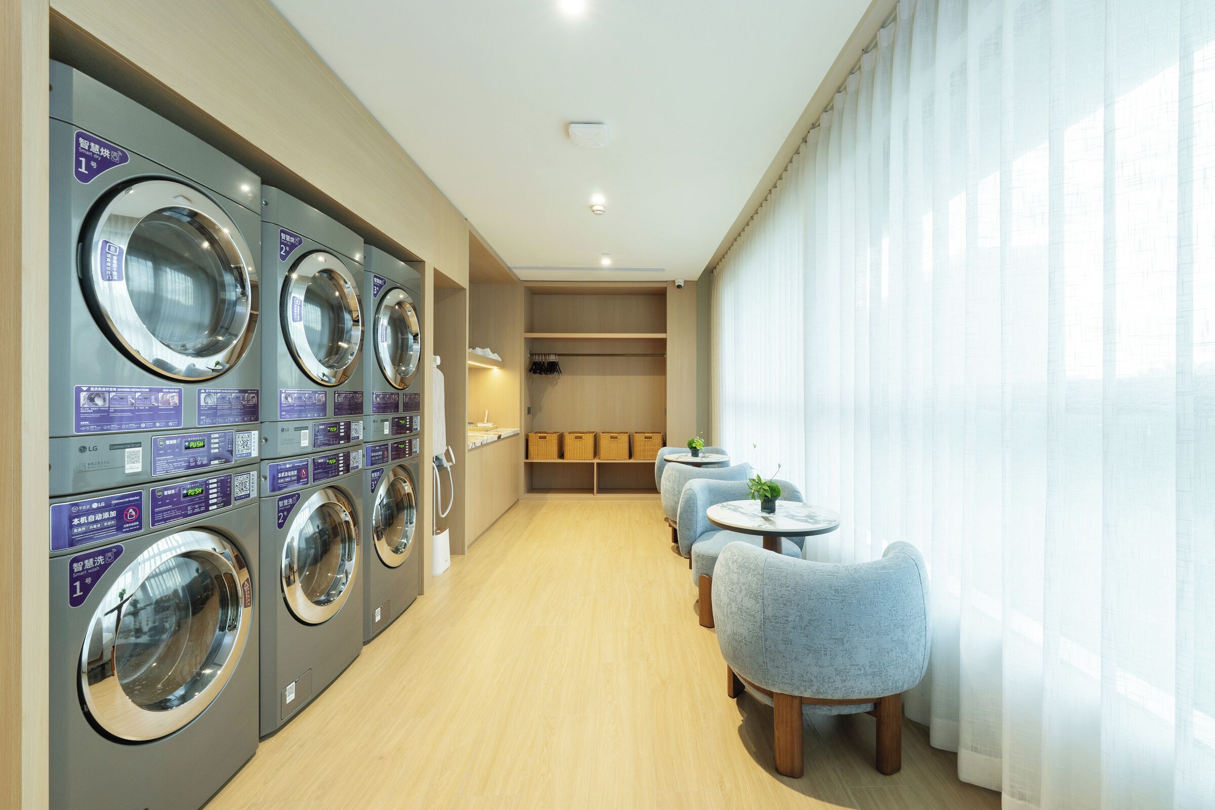 A laundry area with multiple washing machines, a seating area with chairs, and a view of the outside through large windows.
