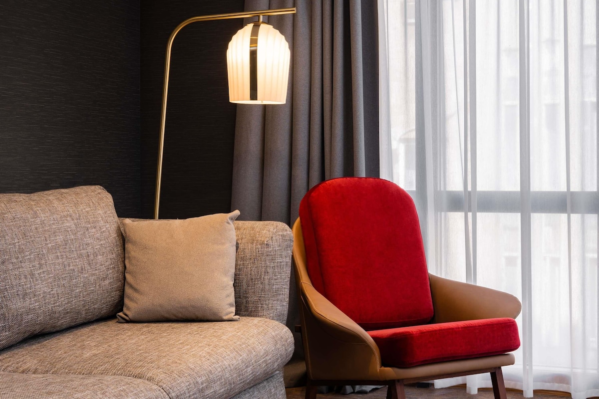Ett modernt vardagsrum med en soffa, en röd fåtölj, en golvlampa och ett fönster med tunna, genomskinliga gardiner.