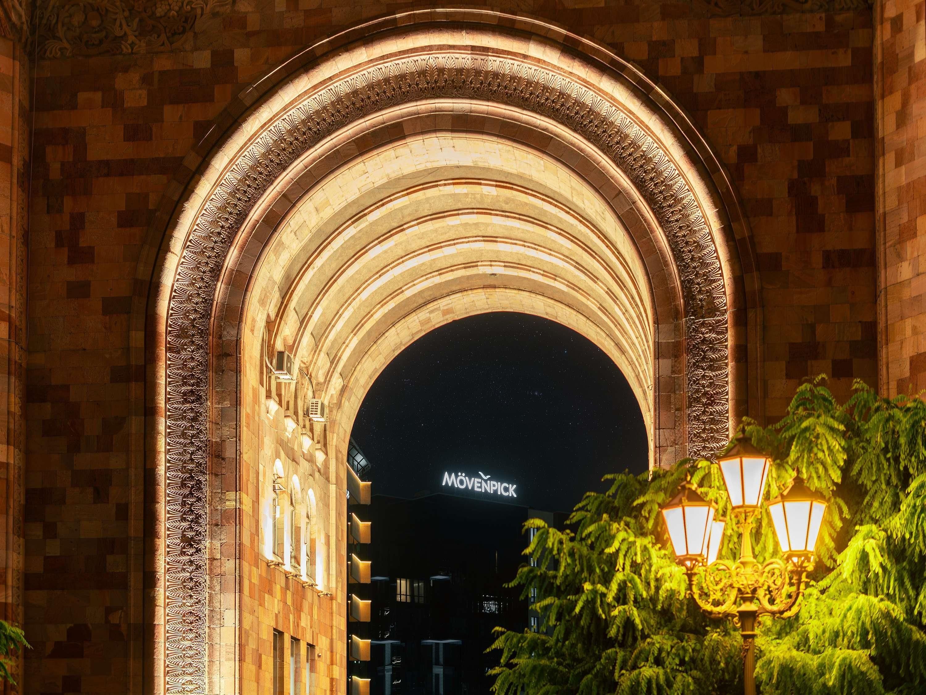 An arched architectural feature with intricate carvings, a sign reading 'MÖVENPICK,' and a street lamp with greenery in the foreground.