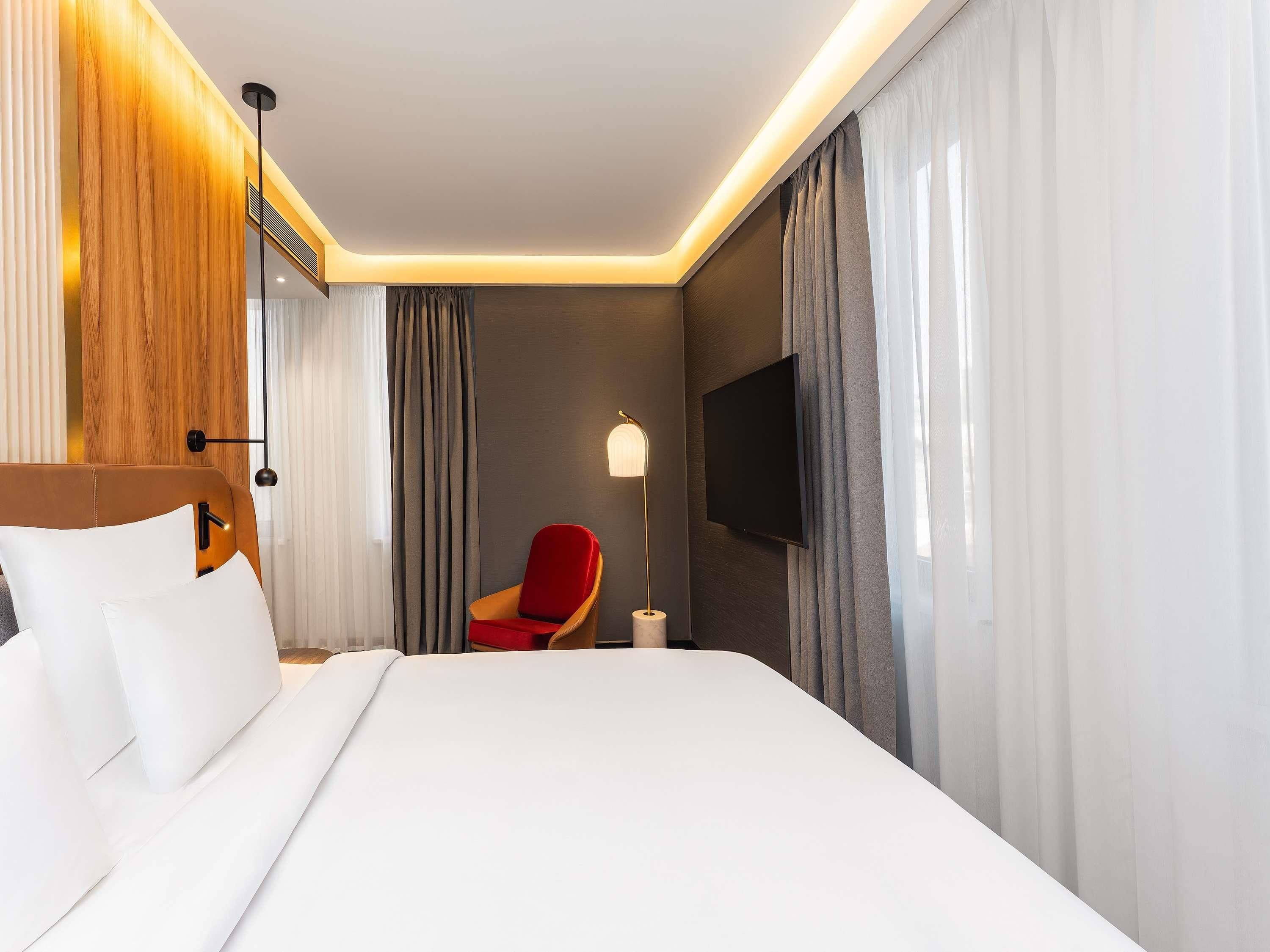 Une chambre d’hôtel avec un lit, un fauteuil rouge, un téléviseur à écran plat et une lampe.