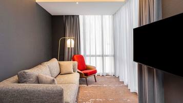 Een moderne hotelkamer met een bank, een rode stoel, een vloerlamp en een groot televisiescherm.