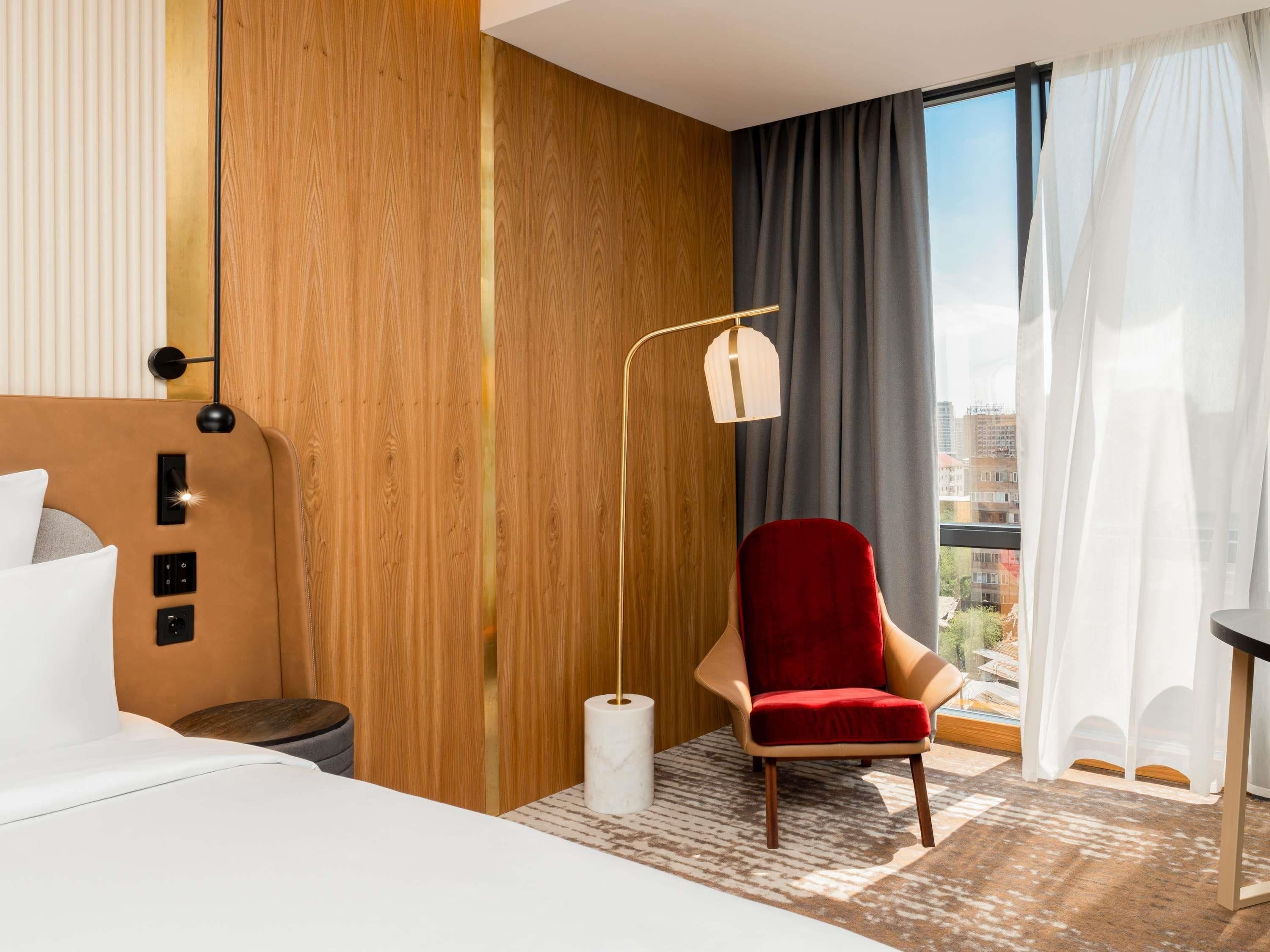 Habitación de hotel moderna con una cama, una silla roja, una lámpara de pie y vistas a la ciudad a través de amplios ventanales.