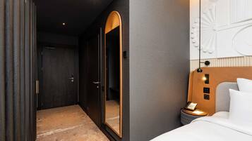 A modern hotel room with a large mirror, a bed with a headboard, a bedside table with a lamp, and a wardrobe.