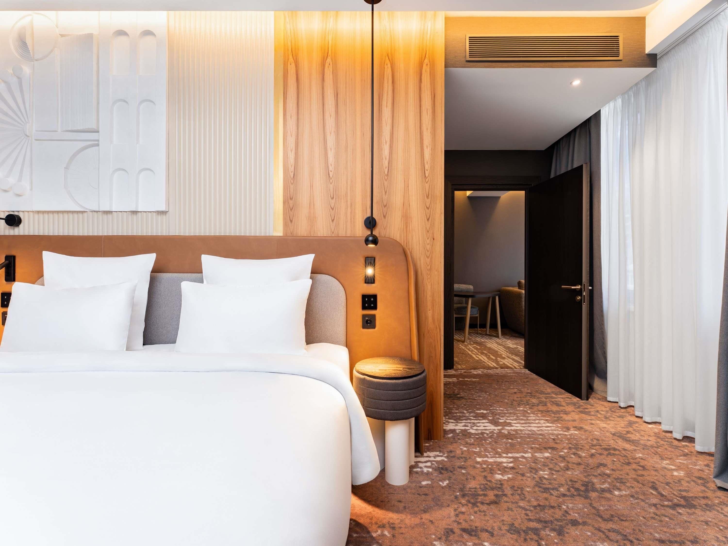 A modern hotel room with a large bed, a bedside table, a lamp, and a view into the adjacent room.