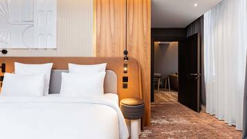 A modern hotel room with a large bed, a bedside table, a lamp, and a view into the adjacent room.